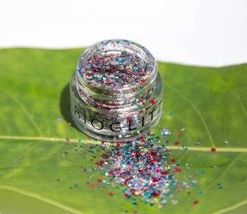 A tiny jar of multicolored glitter sits connected a leaf, with glitter spilling out, suggesting a vibrant, eco-friendly cosmetic product