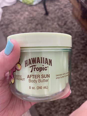 A hand holds a jar of Hawaiian Tropic Exotic Coconut After Sun Body Butter, 8 oz (240 mL). The jar is labeled with details of ingredients