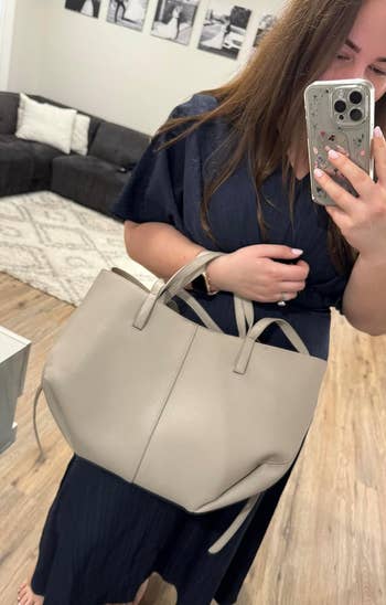 Reviewer in a dark dress holding a light gray tote bag, taking a selfie in a living room