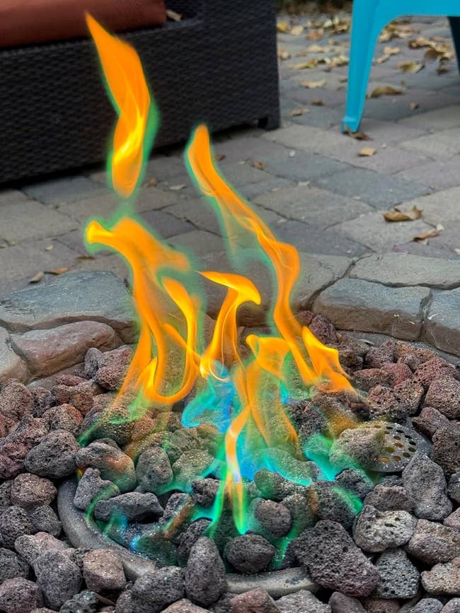 Close-up of a fire pit with vibrant orange and green flames among rocks, placed on a patio with outdoor furniture