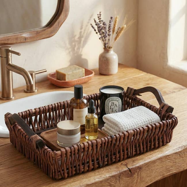 Bathroom countertop with a wicker tray holding skincare bottles, a candle, a folded towel, and a small wooden dish