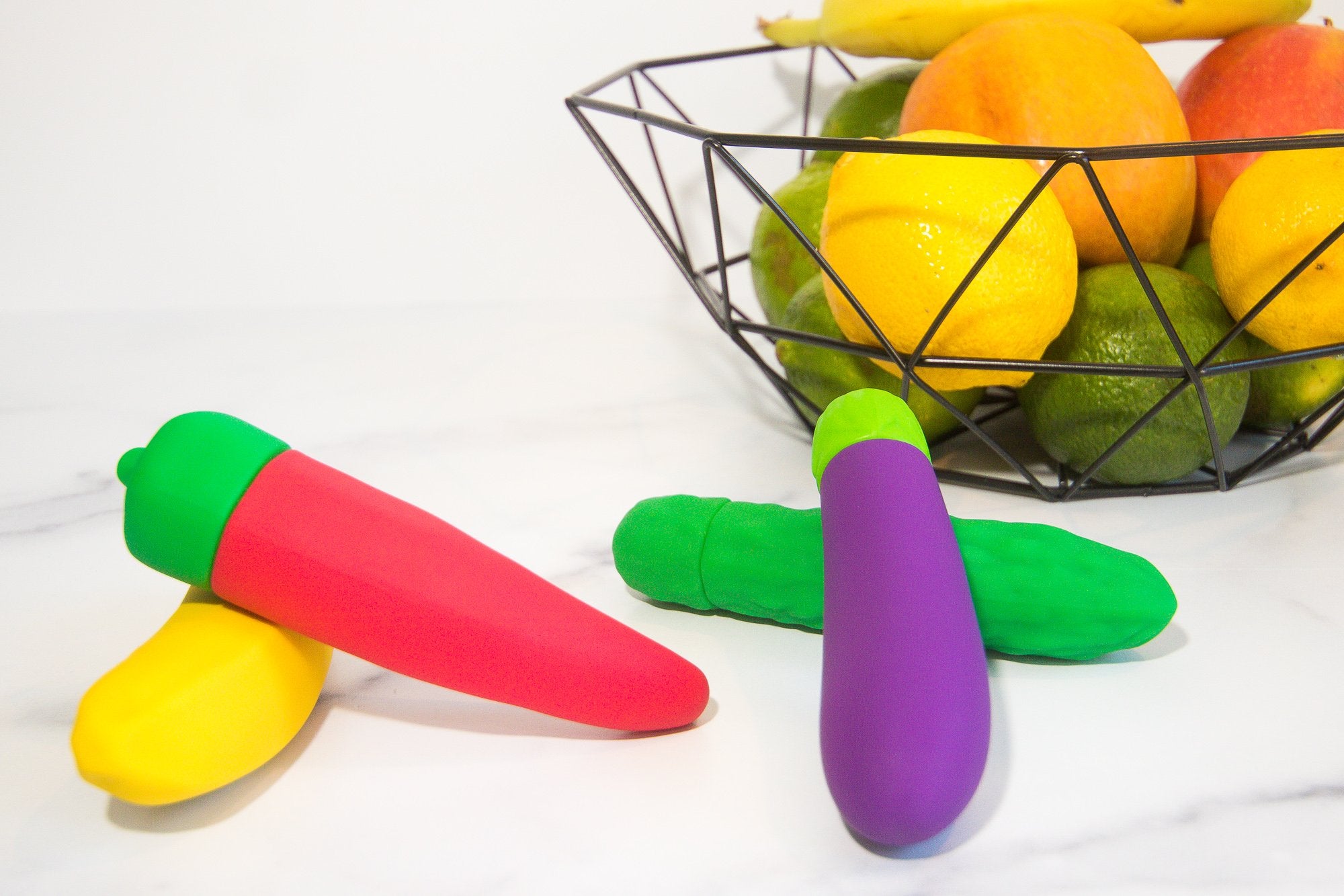 Red chili pepper vibrator, yellow banana vibrator, purple eggplant vibrator and green pickle vibrator next to fruit basket