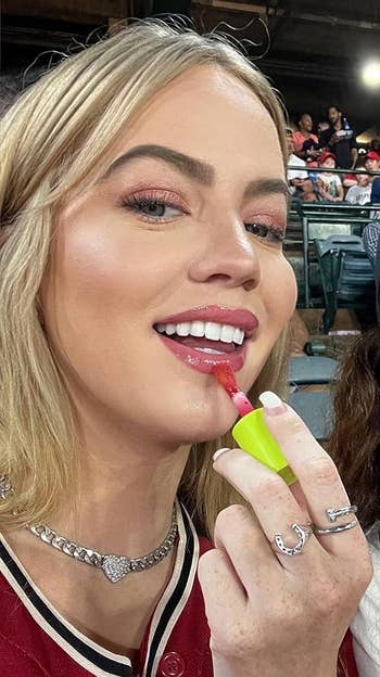 Person smiling, applying articulator  gloss astatine  an outdoor event. Wearing a necklace and rings. Crowd seated successful  the background