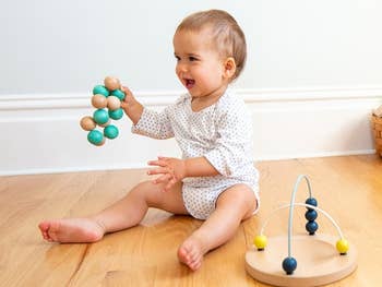 Baby playing with artifact beads connected the floor, holding a woody rattle and sitting adjacent an abacus-like toy