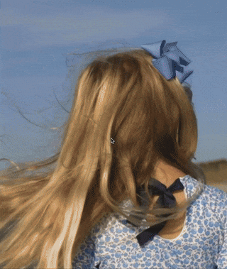 A young girl with long hair wearing a floral-patterned dress and a large bow in her hair looks towards an open field under a clear sky