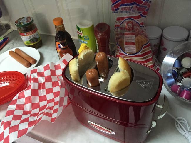 Hot dogs and buns in a toaster with condiments and napkins, ready for a quick meal. Cups and kitchen items on the counter
