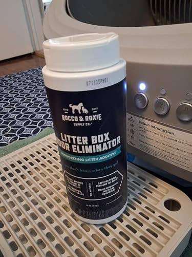 Container of Rocco & Roxie litter box odor eliminator on a tray, beside a litter box appliance with buttons