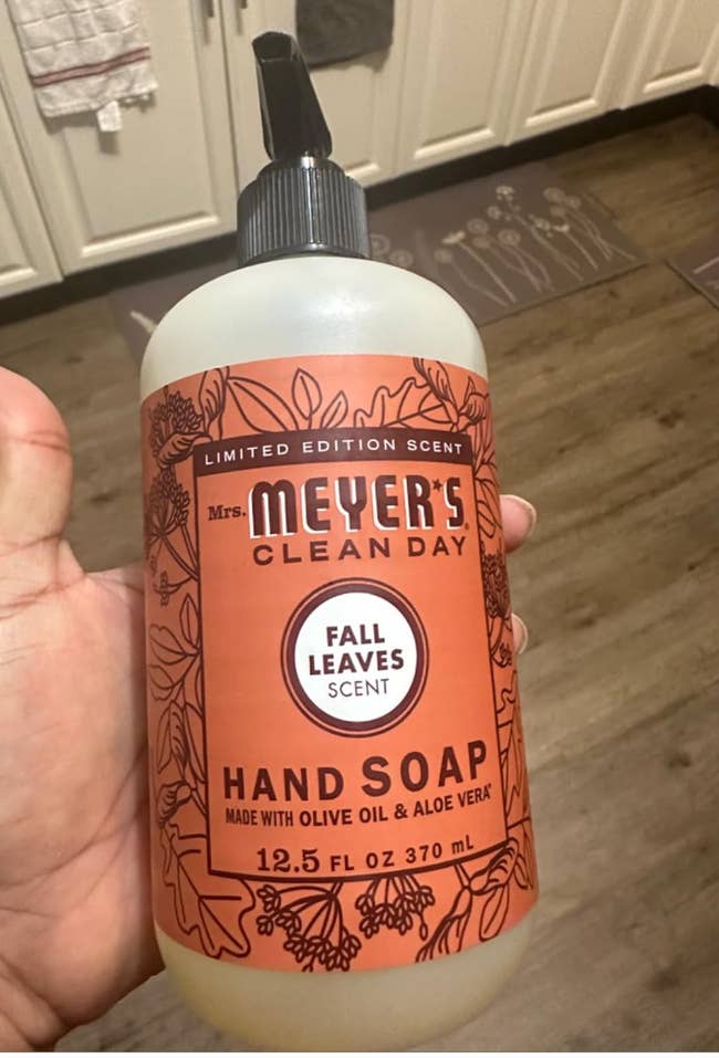 Hand holding a vessel of Mrs. Meyer's Clean Day manus soap, Fall Leaves scent, 12.5 fl oz, with olive lipid and aloe vera