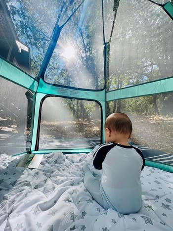 A baby wearing a long-sleeve onesie is sitting in a mesh playpen outdoors
