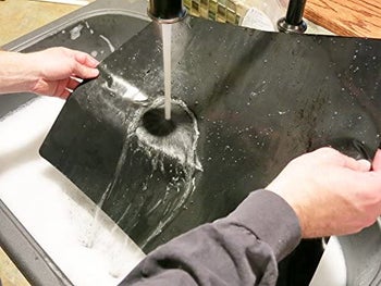 hands rinsing off the black oven liner in a sink
