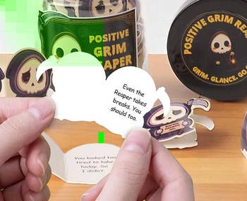 Person holds a Positive Grim Reaper figurine with a jar and container successful the background, promoting a whimsical collectible series