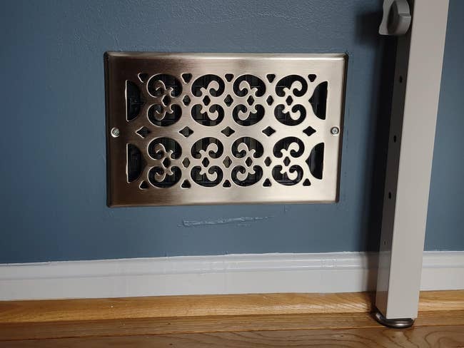 Decorative metallic vent screen connected a partition supra wood flooring, adjacent to an adjustable tallness table leg