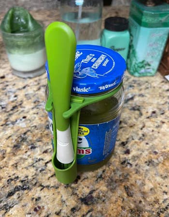 Jar of pickles with a greenish  utensil clipped to the side, holding a pickle spear