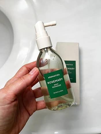 A manus  holds a vessel  of Aromatica Rosemary Root Enhancer. The vessel  is wide   with a pump top, and the statement   is greenish  with achromatic  text