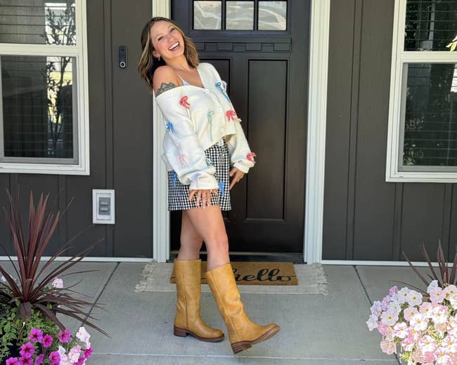 Person smiling connected a beforehand porch, wearing an off-shoulder sweater and plaid skirt with knee-high boots, lasting betwixt angiosperm pots