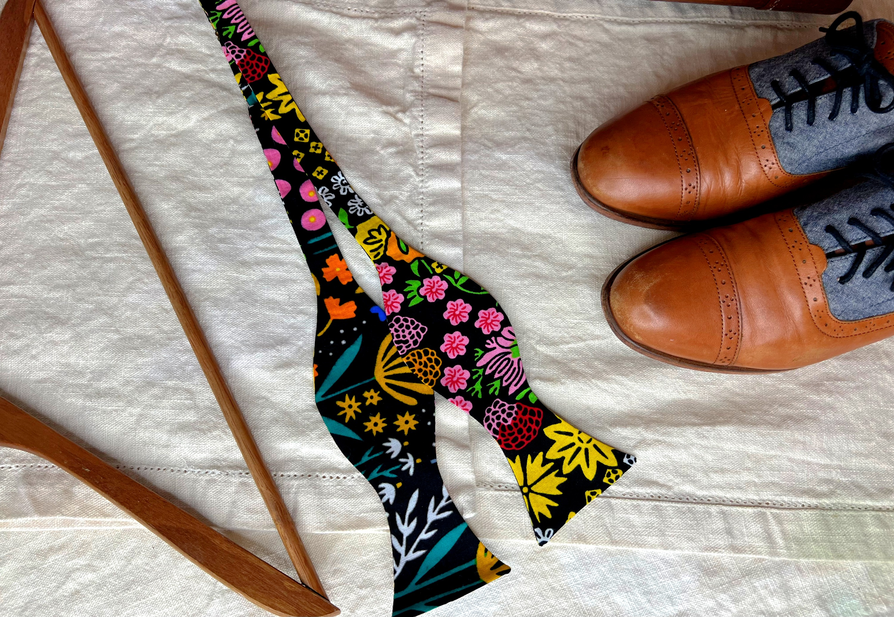 Floral-patterned bow necktie  lies adjacent   a brace  of brownish  formal  shoes and a woody  hanger connected  a cloth surface