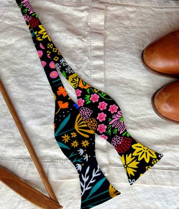Floral-patterned bow necktie lies adjacent a brace of brownish formal shoes and a woody hanger connected a cloth surface