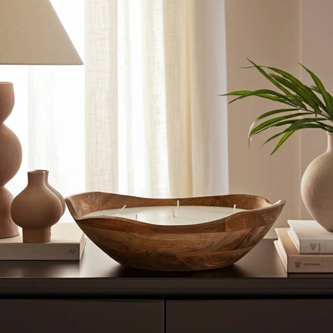 Decorative wooden bowl with wicks, used as a candle, placed on a table with books and a plant, next to a lamp