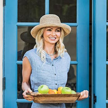 Person in a casual button-up dress and straw hat, holding a wooden tray with apples and oranges, standing by a window