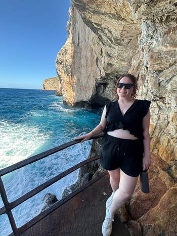 Woman in black ruffled crop top and shorts poses by rocky seaside cliff, holding a water bottle, and smiling at the camera