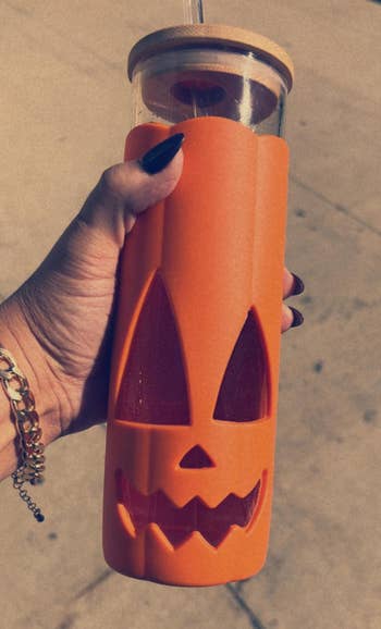 A manus holds a solid vessel with a carved pumpkin design, featuring a bamboo lid and straw, perfect for Halloween-themed drinks
