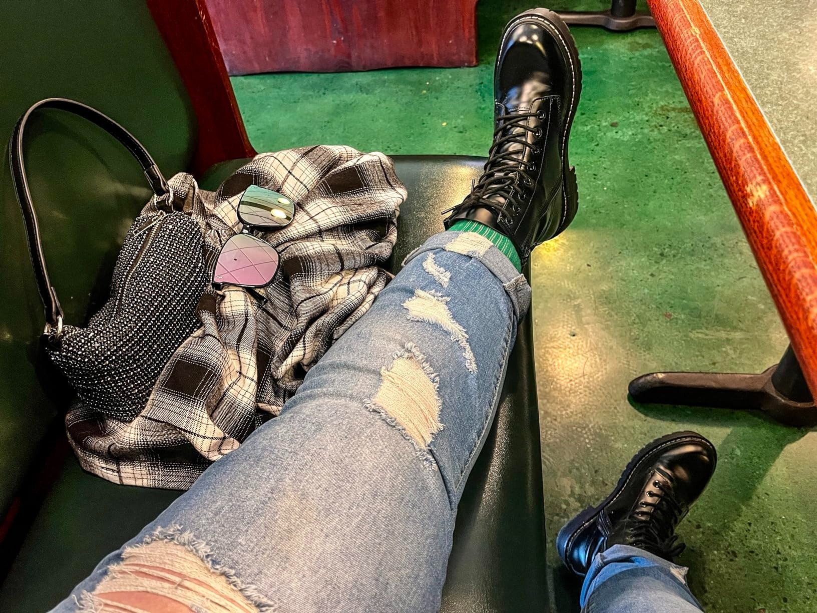 Person wearing ripped jeans and black boots, seated with plaid shirt and studded bag beside them on a green bench