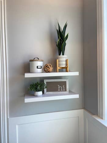 Two white floating shelves with decor items: a plant in a woven pot, a white canister, a twine ball, a small plant, and a sign with