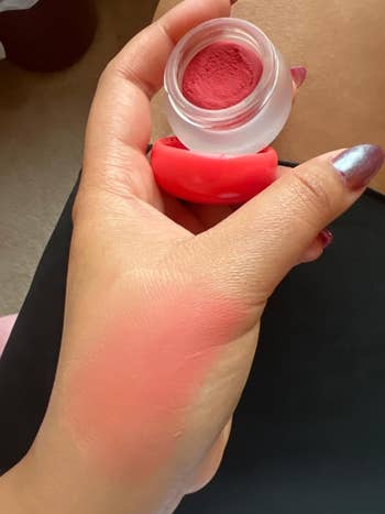 Hand holding an open container of blush, with a swatch on the wrist, showcasing the product's texture and pigment for a shopping article