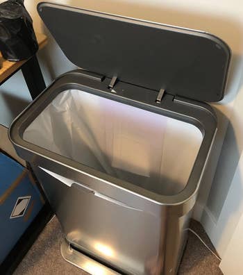 Stainless steel trash can with foot pedal and a raised lid beside a desk
