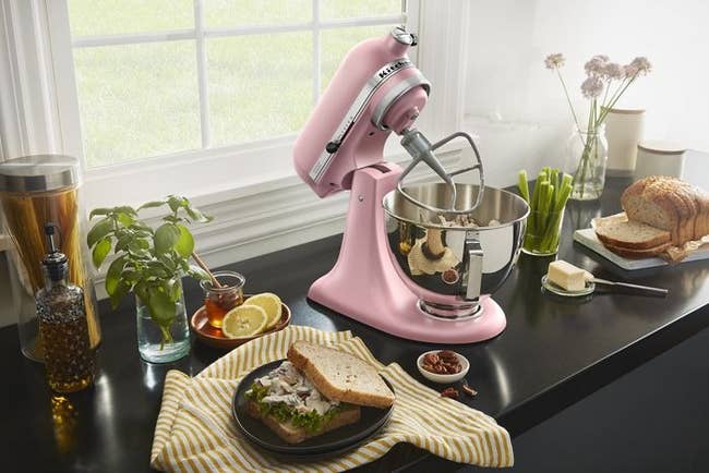 Pink stand mixer on counter with bread, butter, and plants nearby; window view in the background