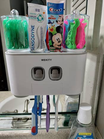 reviewer's white mounted holder with toothbrushes, tubes of toothpaste, and flossing sticks
