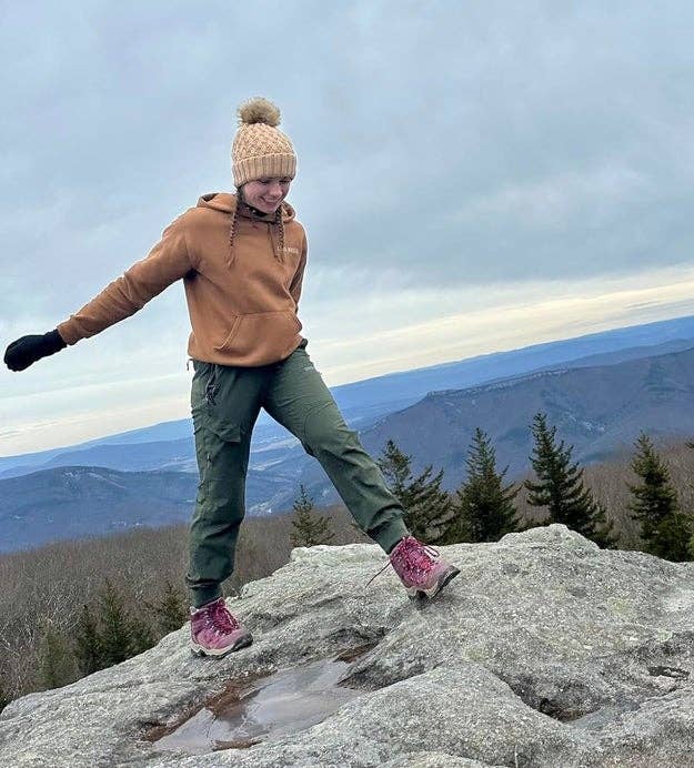 Person in casual outdoor clothing and knit hat stands on a rocky mountain top, enjoying the view of distant mountains