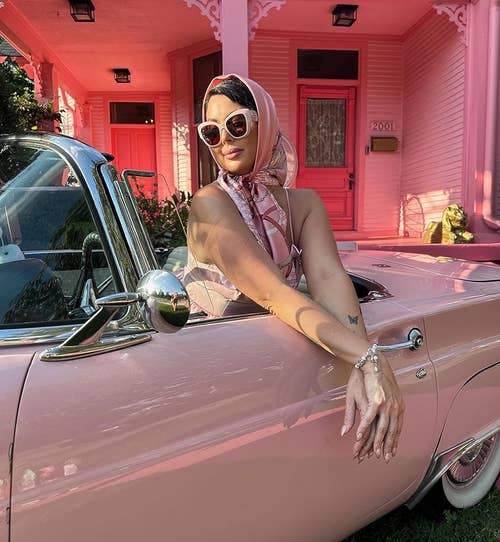 Woman leaning on a vintage car, wearing a scarf, sunglasses, and chic attire. Suitable for an article on fashionable shopping trends
