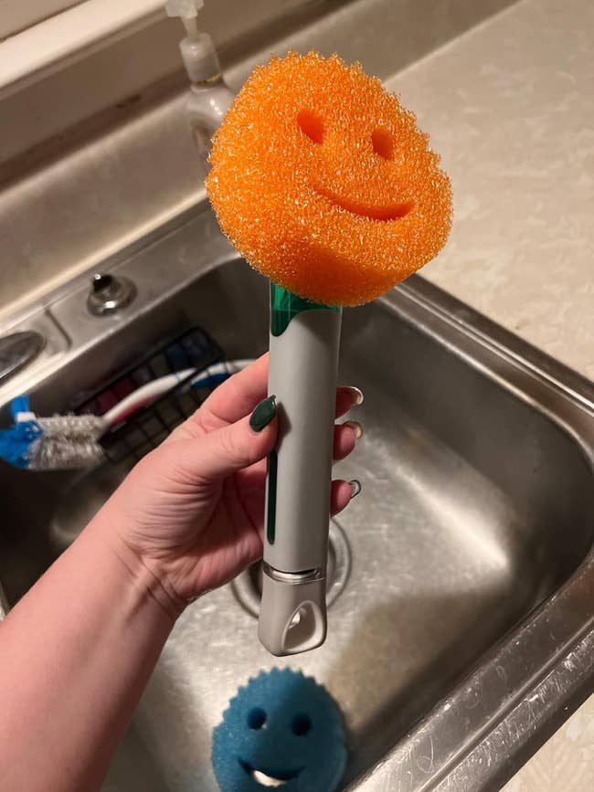 A manus holds a smiling orangish crockery scrubber with grip implicit a sink, showcasing cleaning tools