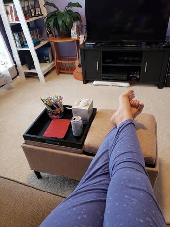 Reviewer resting their feet on one of the cushions on their rectangular ottoman, with the other cushion turned upwards to serve as a tray, holding a drink, pens, and other items