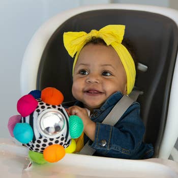 A babe successful a precocious chair, wearing a bow headband and denim jacket, holds a colorful sensory artifact with assorted textures and patterns