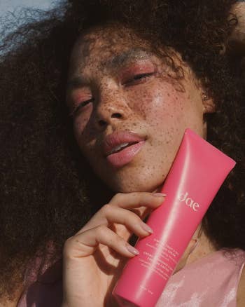 A person with freckles and makeup holds a tube of Dae hair product close to their face. They have voluminous curly hair and are posing outdoors