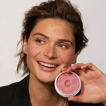 Woman smiling and holding a blush compact adjacent  to her face, showcasing a constitution  merchandise  perfect  for enhancing earthy  beauty