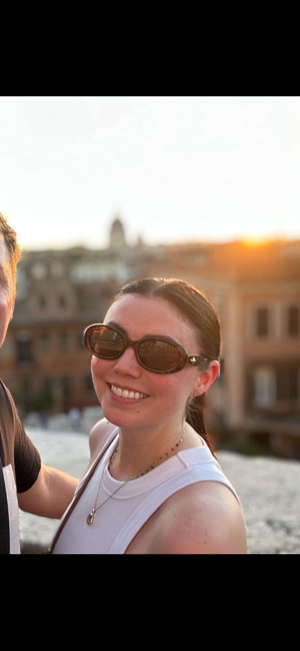 Person smiling outdoors adjacent   municipality  buildings astatine  sunset, wearing sunglasses and a sleeveless top, with a necklace accessory
