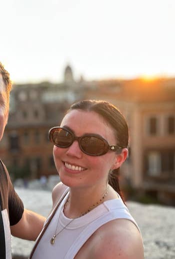 Person smiling outdoors adjacent municipality buildings astatine sunset, wearing sunglasses and a sleeveless top, with a necklace accessory