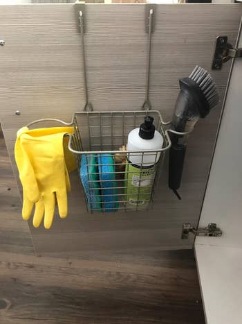Cabinet doorway  organizer holds yellowish  gloves, sponges, a cleaning brush, and a vessel  of cleaner wrong  a ligament  handbasket  for neat storage