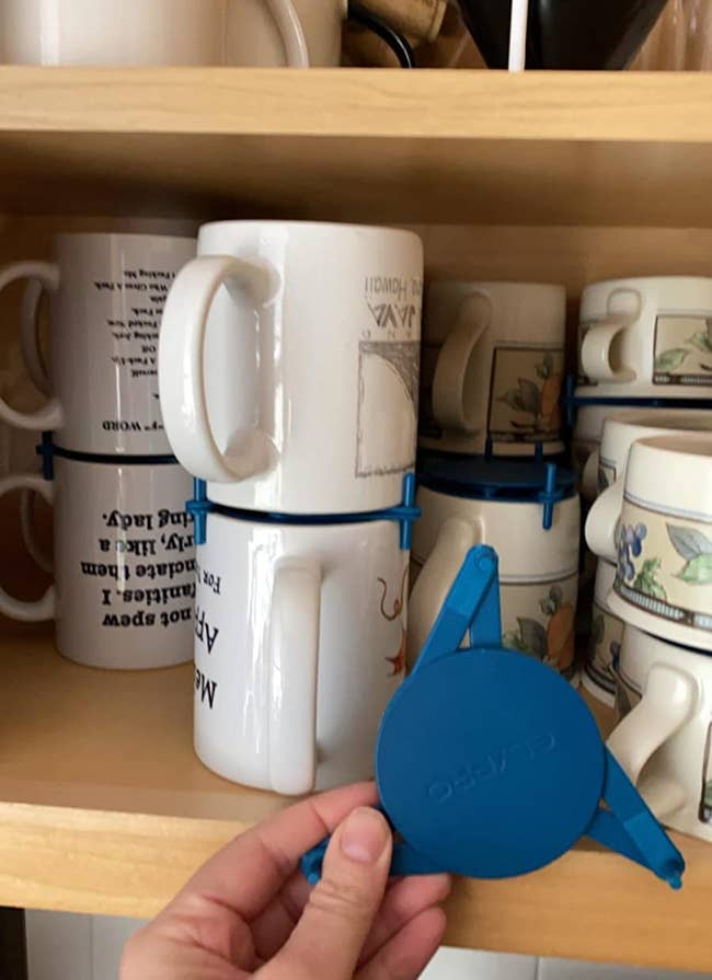 image of reviewer's hand holding blue mug organizer in front of cabinet where mugs are stacked using the same blue organizers