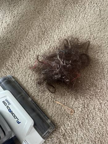 A vacuum cleaner next to a large clump of tangled hair on a carpet, with a comb nearby