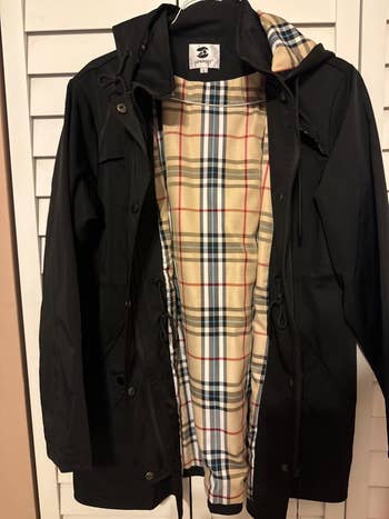 Jacket with a plaid-lined interior hanging connected  a door, showcasing its hood and buttoned front