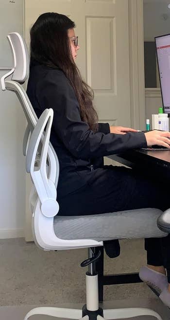 Person in ergonomic chair at desk, working on a computer. Office supplies visible. Casual black outfit