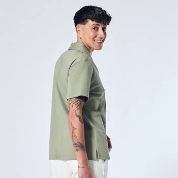 Person smiling, wearing a casual short-sleeve garment and airy pants, showing a elaborate tattoo connected their arm