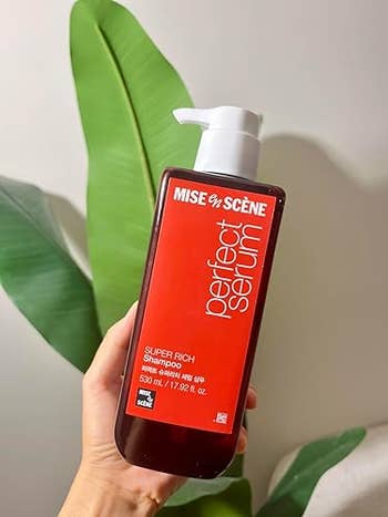 Hand holds a vessel  of Mise En Scène Perfect Serum Super Rich Shampoo, 530ml, against a works  backdrop