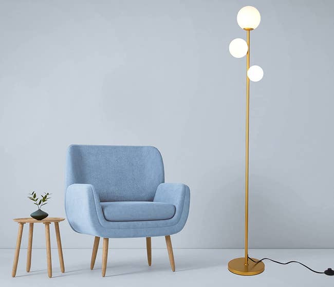Gold skinny lamp with three bulbs and a black cord with a floor push button next to a light blue armchair and wooden side table