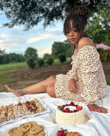Person seated on a picnic blanket with various desserts like cakes and cookies around, wearing a floral outfit