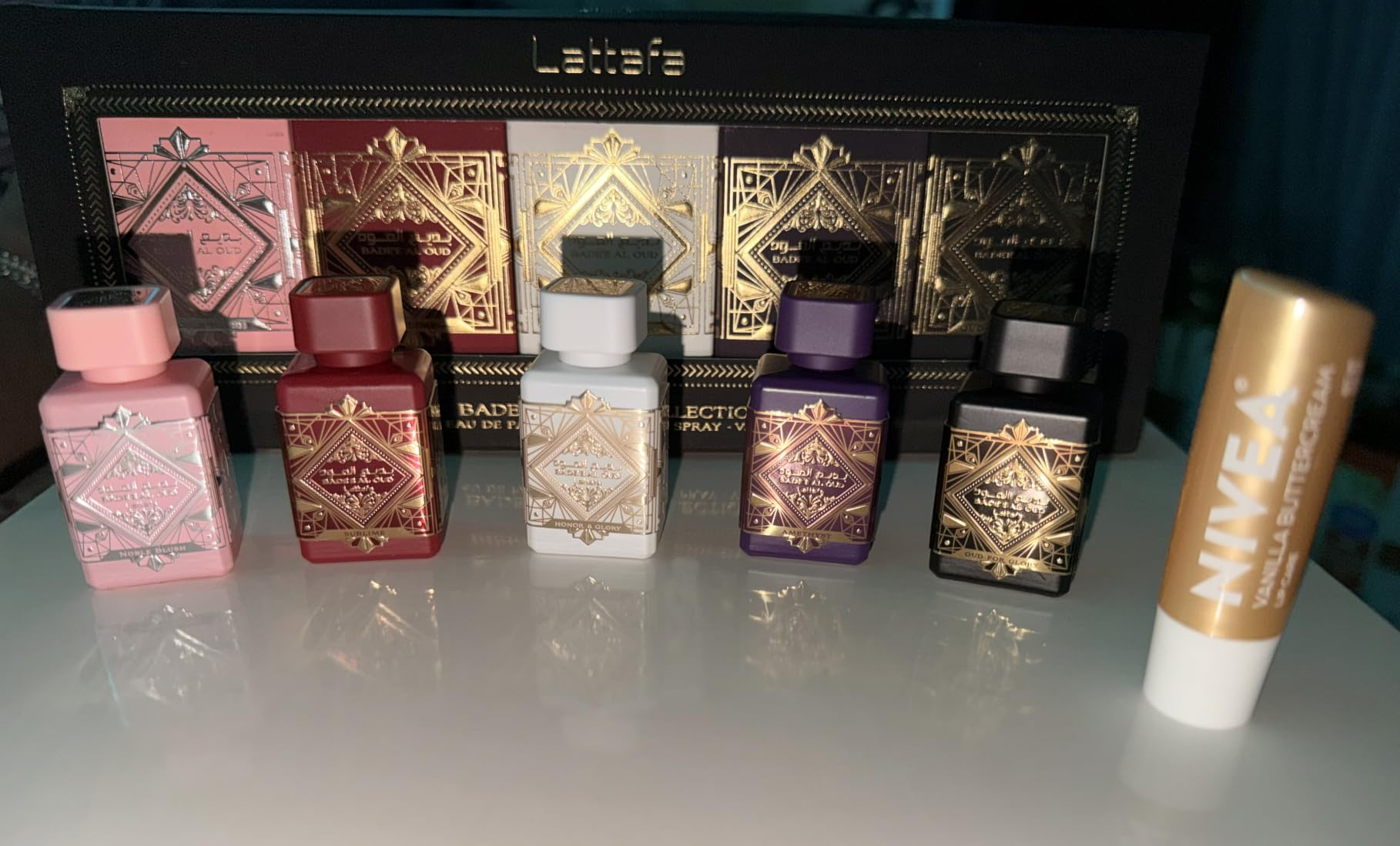 Set of six luxurious perfume bottles with ornate designs, alongside a Nivea articulator  balm, displayed connected  a reflective surface